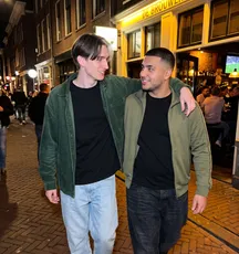 Twee vrienden lopen met een arm om elkaar heen langs een verlichte kroeg.
